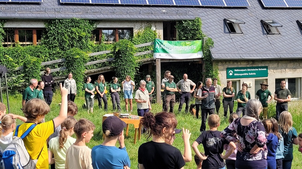 Uwe Borrmeister (m. r.), Leiter der Nationalpark- und Forstverwaltung von Sachsenforst, und Margitta Jendrzejewski (m. l.), Leiterin der Naturbildungsstätte Sellnitz, begrüßen traditionell die Schülerinnen und Schüler, Lehrkräfte sowie Mitarbeitende und Freiwillige der Nationalpark- und Forstverwaltung zu den Waldjugendspielen in der Nationalparkregion Sächsische Schweiz. Das Foto stammt von 2025; hier ist Laura Larissa Lepczynski (h. 2. v. l.) als Freiwillige eingebunden.  Foto: Hp. Mayr